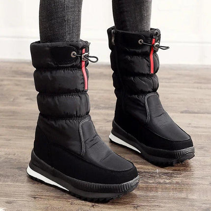 Bota negra para nieve impermeable de piel sintética para mujer, con cierre rojo, en interior de madera.